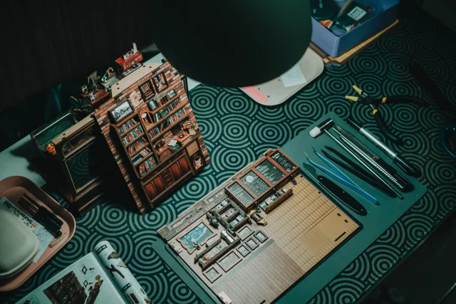 A detailed miniature bookshelf crafting setup with tools on a patterned table. Fitting a book nook kit workspace into one shared room