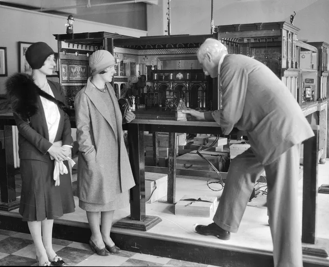 At left, 2 women (Katherine L. Howes[?] and Mrs. Franklin A.[?] Howe, Jr.) stand in front of Sir Nevile Wilkinson's "Titania's Palace." Both women face toward Sir Wilkinson at right. Sir Wilkinson stands in profile, facing left. He leans forward and slightly onto the table supporting the miniature p. Miniature room ideas: the ones with staying power, 2027 edition